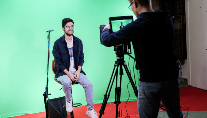 Ein junger Mann wird vor einem von einem Kameramann vor einem Greenscreen gefilmt. Er sitzt auf einem höhenverstellbaren Barhocker und seitlich rechts vor ihm ist ein kleines Richtmikrofon aufgebaut.