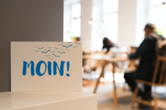 Postkarte mit der Aufschrift „Moin!“ vor Menschen an Leseplätzen in einer Bücherhalle
