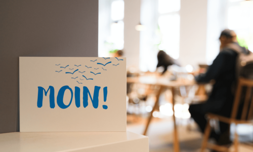 Postkarte mit der Aufschrift „Moin!“ vor Menschen an Leseplätzen in einer Bücherhalle
