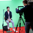 Ein junger Mann wird vor einem von einem Kameramann vor einem Greenscreen gefilmt. Er sitzt auf einem höhenverstellbaren Barhocker und seitlich rechts vor ihm ist ein kleines Richtmikrofon aufgebaut.