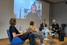 Vier Personen sitzen auf einem kleinen Podium und lächeln einander freundlich an. Hinter ihnen an der Wand eine Projektion einer Diskussionsteilnehmerin.