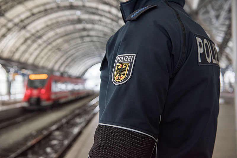 Foto-Bildausschnitt zeigt die Jacke eines Polizisten, der am Bahnhof gegenüber eines Zuges steht
